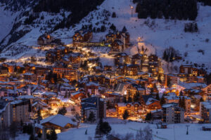 A snow-covered ski resort Les Deux Alpes illuminates as golden lights shine from chalet windows, reflecting off the white slopes under a fading sky